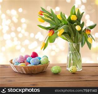 easter, holidays and object concept - colored eggs in wicker basket and tulip flowers in vase on wooden table over festive lights on beige background. colored easter eggs in basket and tulip flowers