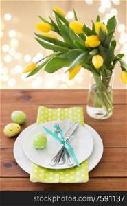 easter, holidays and object concept - green colored egg, plates with cutlery and tulip flowers in vase on wooden table over festive lights on beige background. easter egg, plates, cutlery and tulip flowers