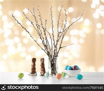 easter, holidays and object concept - pussy willow branches, colored eggs and chocolate bunnies on table over festive lights background. pussy willow, easter eggs and chocolate bunnies