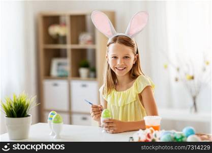 easter, holidays and people concept - happy girl wearing bunny ears headband coloring eggs with colors and brush at home. happy girl coloring easter eggs at home