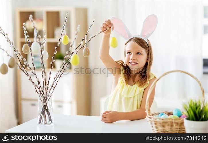easter, holidays and people concept - happy girl wearing bunny ears headband decorating willow branches by toy eggs at home. girl decorating willow by easter eggs at home
