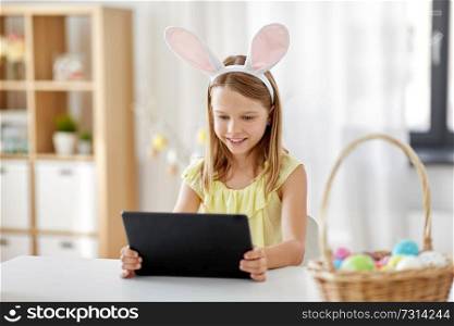 easter, holidays and people concept - happy girl wearing bunny ears headband with tablet pc computer and basket of colored eggs at home. happy girl with tablet pc and easter eggs at home