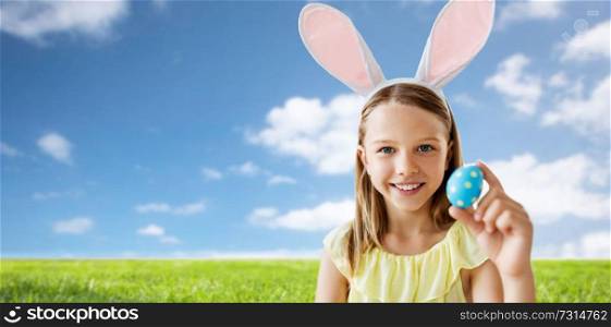 easter, holidays and people concept - happy girl wearing bunny ears headband with colored egg over blue sky and grass background. happy girl with colored easter egg