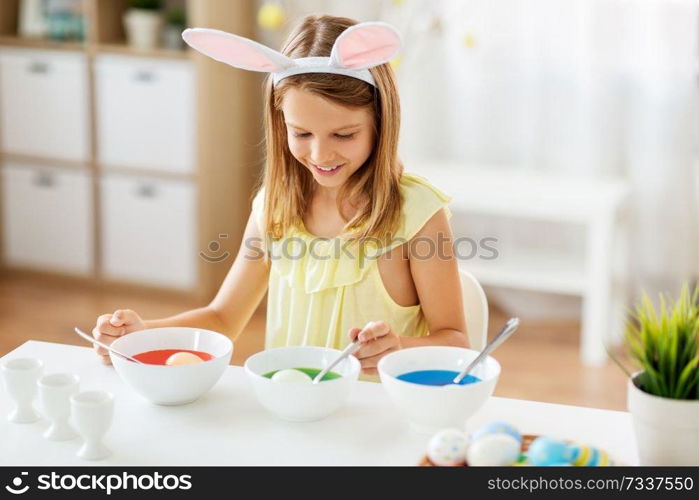 easter, holidays and people concept - happy girl wearing bunny ears headband coloring eggs by liquid dye at home. girl coloring easter eggs by liquid dye at home