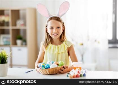easter, holidays and people concept - happy girl wearing bunny ears headband with colored eggs at home. happy girl with colored easter eggs at home