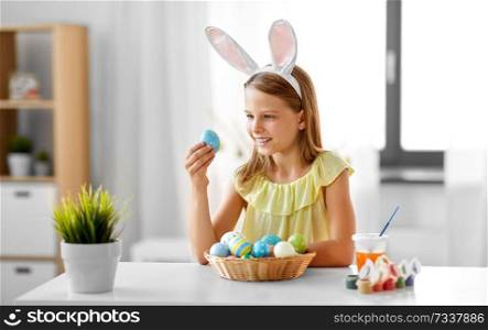 easter, holidays and people concept - happy girl wearing bunny ears headband with colored eggs at home. happy girl with colored easter eggs at home