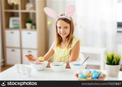 easter, holidays and people concept - happy girl wearing bunny ears headband coloring eggs by liquid dye at home. girl coloring easter eggs by liquid dye at home