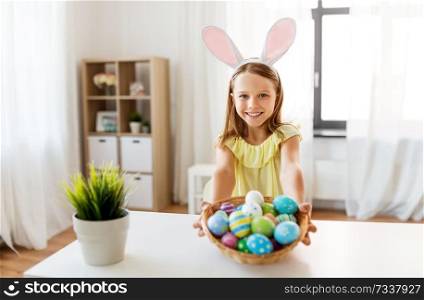 easter, holidays and people concept - happy girl wearing bunny ears headband with basket of colored eggs at home. happy girl with colored easter eggs at home