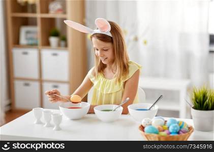 easter, holidays and people concept - happy girl wearing bunny ears headband coloring eggs by liquid dye at home. girl coloring easter eggs by liquid dye at home