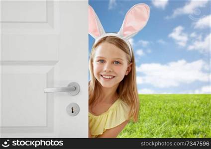 easter, holidays and people concept - happy girl wearing bunny ears peeking out door over blue sky and grass background. happy girl with easter bunny ears peeking out door