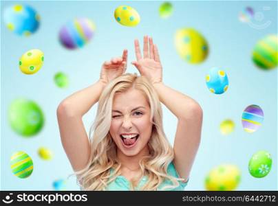 easter, holidays and people concept - happy smiling young woman making bunny ears over blue background with colored eggs. happy woman making bunny ears over easter eggs