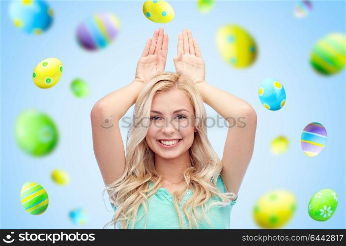easter, holidays and people concept - happy smiling young woman making bunny ears over blue background with colored eggs. happy woman making bunny ears over easter eggs
