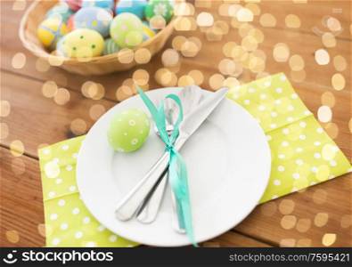 easter, holidays and table setting concept - close up of plate with cutlery and colored eggs on wooden table. close up of table setting and easter eggs