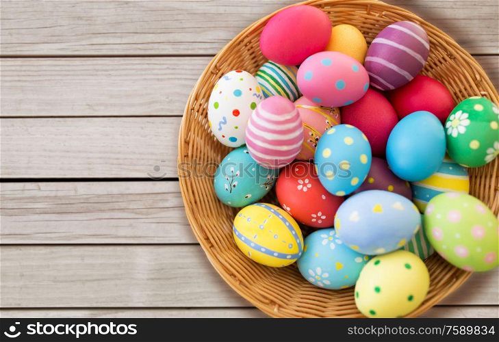 easter, holidays and tradition concept - close up of colored eggs in wicker basket on grey wooden boards background. close up of colored easter eggs in basket