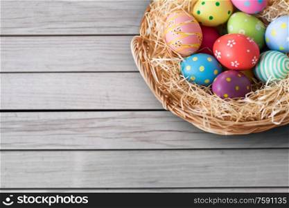 easter, holidays and traditions concept - close up of colored eggs in wicker basket over grey wooden boards background. close up of colored easter eggs in basket
