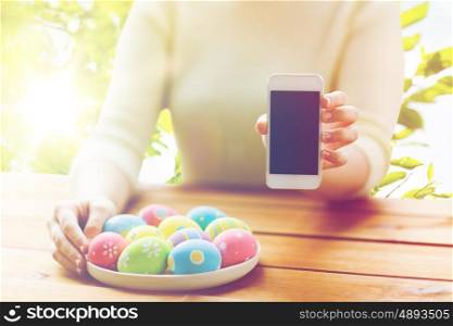easter, holidays, technology and people concept - close up of woman hands with colored easter eggs on plate and smartphone