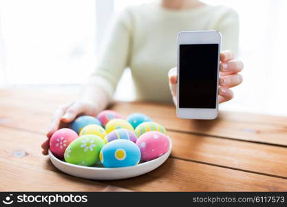 easter, holidays, technology and people concept - close up of woman hands with colored easter eggs on plate and smartphone