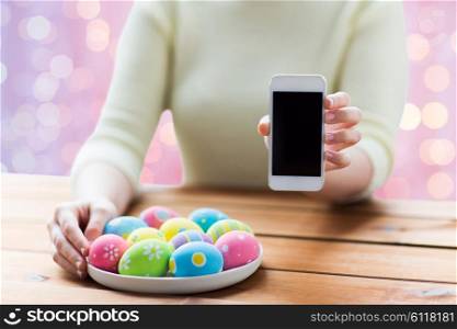 easter, holidays, technology and people concept - close up of woman hands with colored easter eggs on plate and smartphone