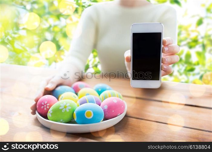easter, holidays, technology and people concept - close up of woman hands with colored easter eggs on plate and smartphone over green natural background