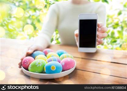 easter, holidays, technology and people concept - close up of woman hands with colored easter eggs on plate and smartphone over green natural background