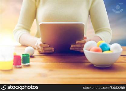 easter, holidays, technology and people concept - close up of woman hands with tablet pc computer, eggs and colors at wooden table over sky background. close up of woman with tablet pc and easter eggs