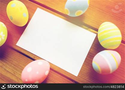 easter, holidays, tradition and object concept - close up of colored easter eggs and blank white paper card on wooden surface with copy space