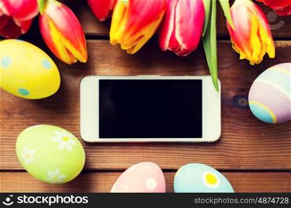 easter, holidays, tradition and object concept - close up of colored easter eggs, tulip flowers and smartphone with blank screen on wooden surface