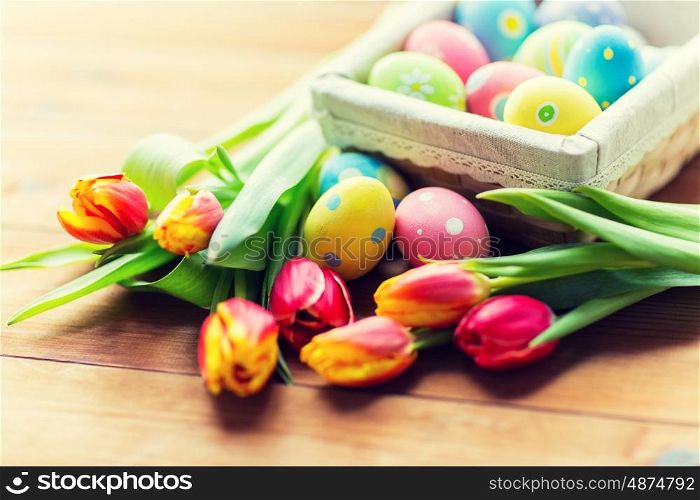 Easter, holidays, tradition and object concept - close up of colored easter eggs in basket and tulip flowers