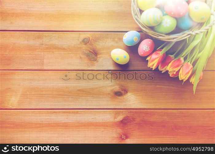easter, holidays, tradition and object concept - close up of colored easter eggs in basket and tulip flowers on wooden table with copy space. close up of easter eggs in basket and flowers