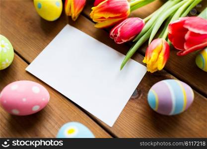 easter, holidays, tradition and object concept - close up of colored easter eggs, tulip flowers and blank white paper card on wooden surface with copy space
