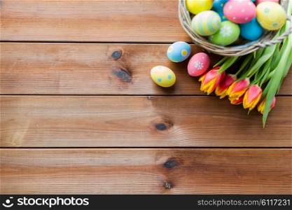 easter, holidays, tradition and object concept - close up of colored easter eggs in basket and tulip flowers on wooden table with copy space