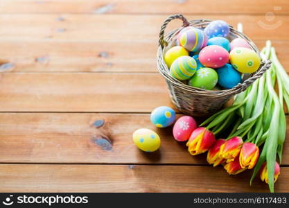 easter, holidays, tradition and object concept - close up of colored easter eggs in basket and tulip flowers on wooden table with copy space