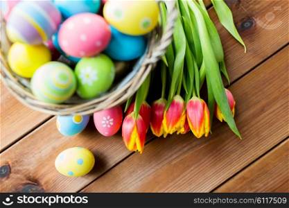 easter, holidays, tradition and object concept - close up of colored easter eggs in basket and tulip flowers on wooden table with copy space