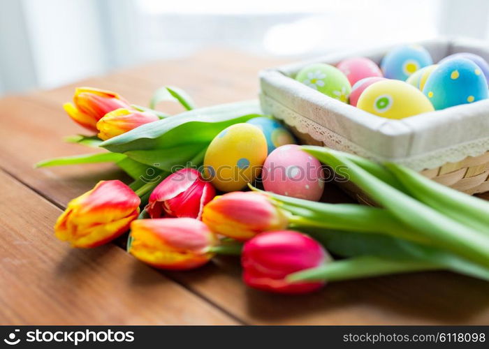 easter, holidays, tradition and object concept - close up of colored easter eggs in basket and tulip flowers