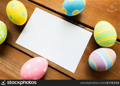 easter, holidays, tradition and object concept - close up of colored easter eggs and blank white paper card on wooden surface with copy space