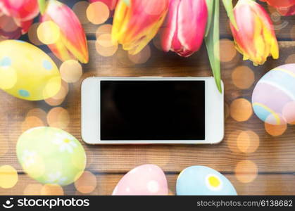 easter, holidays, tradition and object concept - close up of colored easter eggs, tulip flowers and smartphone with blank screen on wooden surface