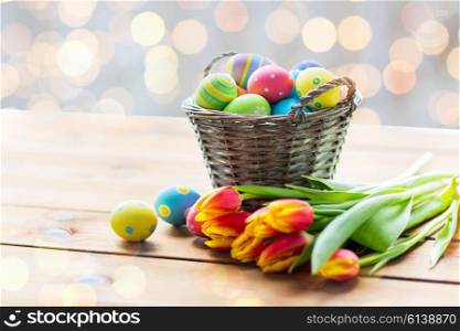 easter, holidays, tradition and object concept - close up of colored easter eggs in basket and tulip flowers on wooden table