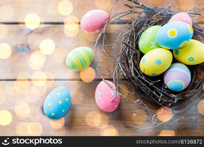 easter, holidays, tradition and object concept - close up of colored easter eggs in nest on wooden surface over holidays lights