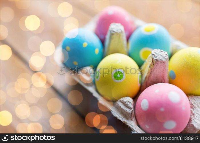 easter, holidays, tradition and object concept - close up of colored easter eggs in egg box or carton wooden surface over holidays lights