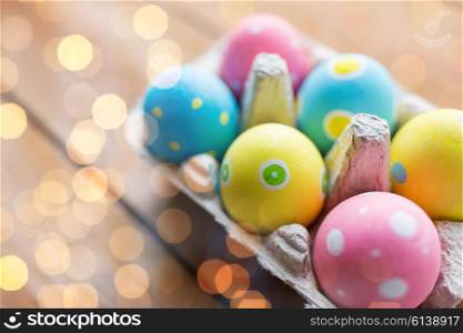easter, holidays, tradition and object concept - close up of colored easter eggs in egg box or carton wooden surface over holidays lights