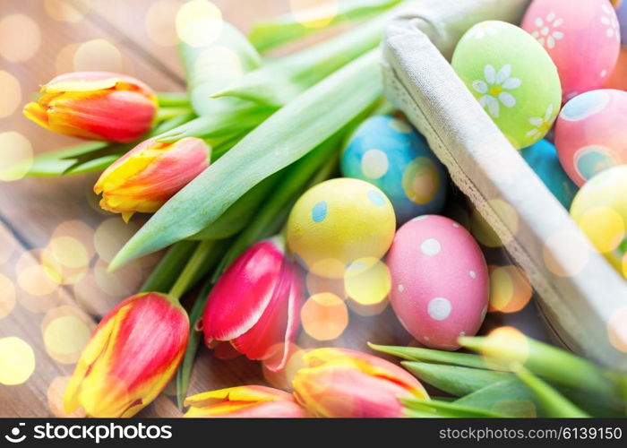 easter, holidays, tradition and object concept - close up of colored easter eggs in basket and tulip flowers over holidays lights