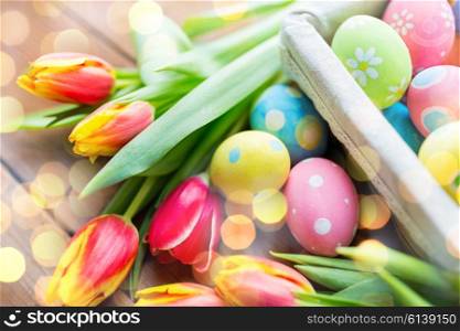 easter, holidays, tradition and object concept - close up of colored easter eggs in basket and tulip flowers over holidays lights