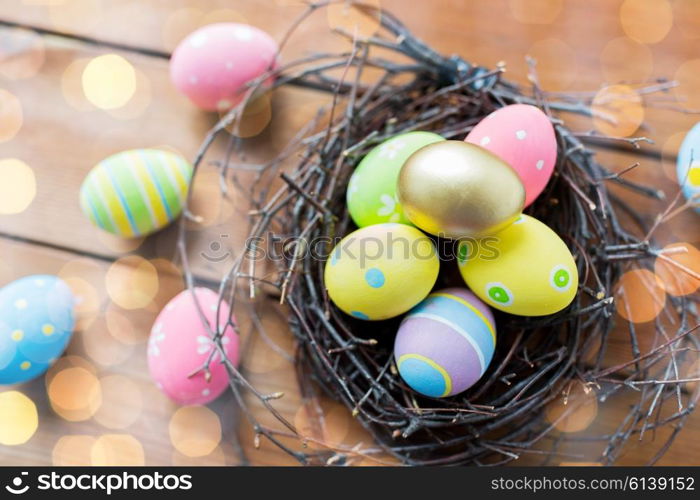 easter, holidays, tradition and object concept - close up of colored easter eggs in nest on wooden surface over lights