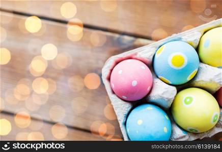 easter, holidays, tradition and object concept - close up of colored easter eggs in egg box or carton wooden surface with copy space over lights