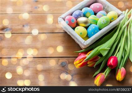 easter, holidays, tradition and object concept - close up of colored easter eggs in basket and tulip flowers over lights