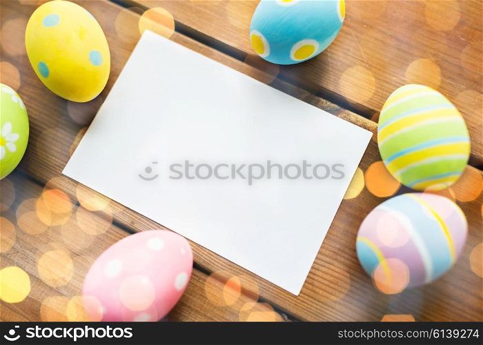 easter, holidays, tradition and object concept - close up of colored easter eggs and blank white paper card on wooden surface with copy space over lights