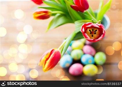 easter, holidays, tradition and object concept - close up of colored easter eggs and tulip flowers in bucket on wooden table over holidays lights