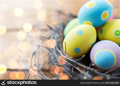 easter, holidays, tradition and object concept - close up of colored easter eggs in nest on wooden surface over holidays lights