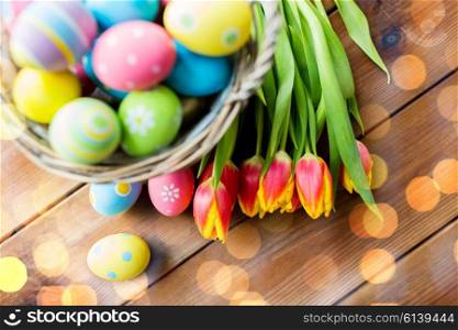 easter, holidays, tradition and object concept - close up of colored easter eggs in basket and tulip flowers on wooden table over holidays lights