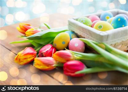 easter, holidays, tradition and object concept - close up of colored easter eggs in basket and tulip flowers over lights